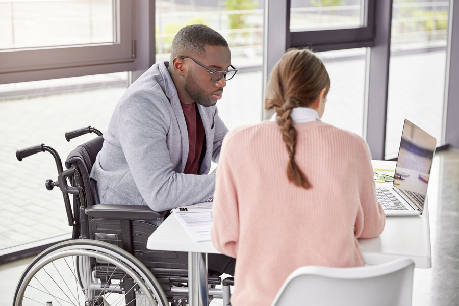 A man in a wheelchair and a woman are using a laptop at a table